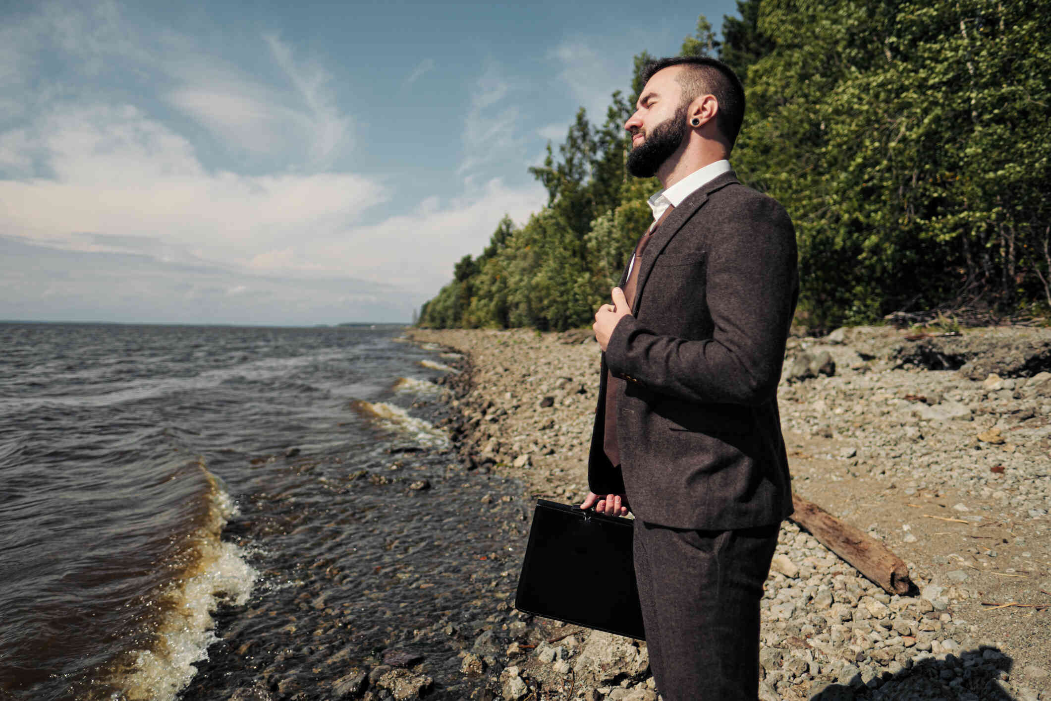 A man in a buisness suit and briefcase stands near the ocean and gazes out while deep in thought.